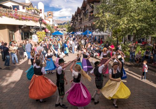 A lively street festival with people in colorful skirts dancing in a circle as crowds stroll along a shaded, festive avenue.