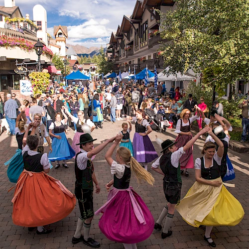 A lively street festival with people in colorful skirts dancing in a circle as crowds stroll along a shaded, festive avenue.