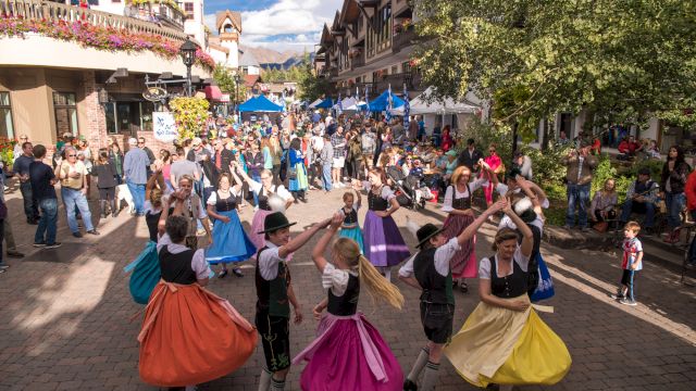 A lively street festival with people in colorful skirts dancing in a circle as crowds stroll along a shaded, festive avenue.