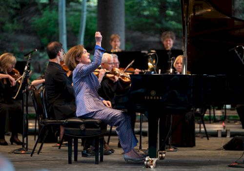 A conductor in a blue suit gestures dramatically at a grand piano as a jazz-like orchestra performs on stage.