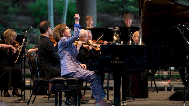 A conductor in a blue suit gestures dramatically at a grand piano as a jazz-like orchestra performs on stage.