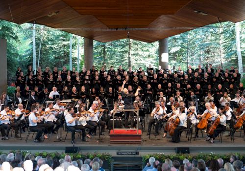 A large choir with an orchestra performing on an outdoor stage, surrounded by trees and an audience watching, all under a wooden pavilion.