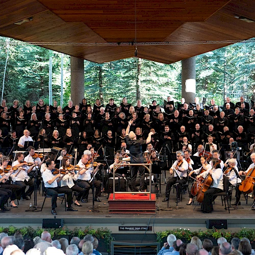 A large choir with an orchestra performing on an outdoor stage, surrounded by trees and an audience watching, all under a wooden pavilion.