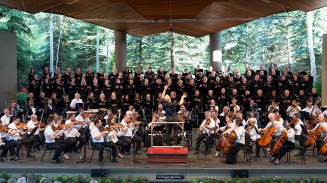 A large choir with an orchestra performing on an outdoor stage, surrounded by trees and an audience watching, all under a wooden pavilion.
