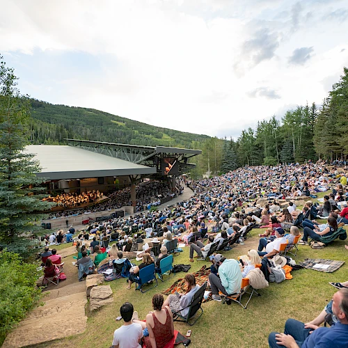 A large outdoor concert with a stage nestled in a forested hillside, and a huge crowd seated on blankets and chairs enjoying the show.