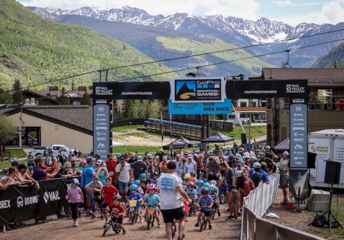 Crowd at a mountain bike race start, banners, riders with helmets, event tents, and a scenic alpine backdrop.