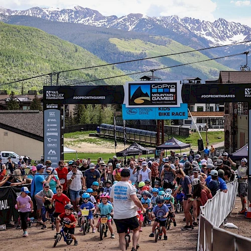 Crowd at a mountain bike race start, banners, riders with helmets, event tents, and a scenic alpine backdrop.