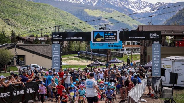 Crowd at a mountain bike race start, banners, riders with helmets, event tents, and a scenic alpine backdrop.