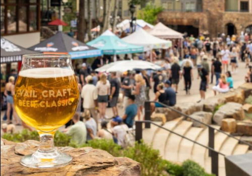 A glass of beer in the foreground at a lively outdoor festival with tents and crowds in the background.