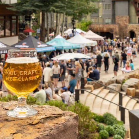 A glass of beer in the foreground at a lively outdoor festival with tents and crowds in the background.