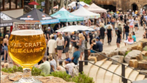 A glass of beer in the foreground at a lively outdoor festival with tents and crowds in the background.