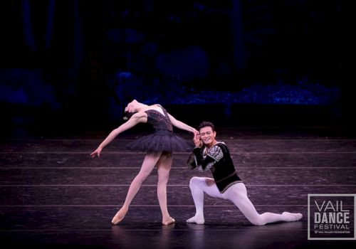 A pair of ballet dancers perform on stage, the female in a dark tutu arching gracefully while the male supports her in a kneeling pose; Vail Dance Festival logo visible in the corner.