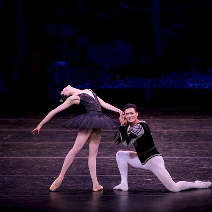 A pair of ballet dancers perform on stage, the female in a dark tutu arching gracefully while the male supports her in a kneeling pose; Vail Dance Festival logo visible in the corner.
