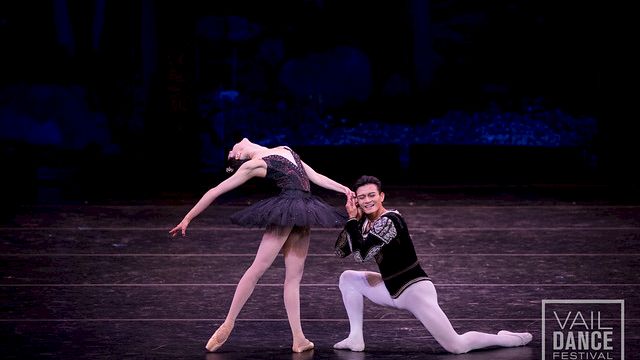 A pair of ballet dancers perform on stage, the female in a dark tutu arching gracefully while the male supports her in a kneeling pose; Vail Dance Festival logo visible in the corner.