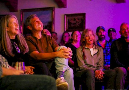 A group of people watching a show or performance in a cozy, dimly lit room, smiling and laughing together.