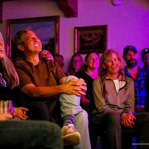 A group of people watching a show or performance in a cozy, dimly lit room, smiling and laughing together.