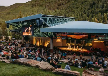 Outdoor amphitheater or cinema in a mountain setting, with a crowd seated on grass and rocks, a stage and screens under a blue pavilion.