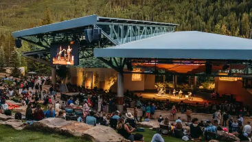 Outdoor amphitheater or cinema in a mountain setting, with a crowd seated on grass and rocks, a stage and screens under a blue pavilion.