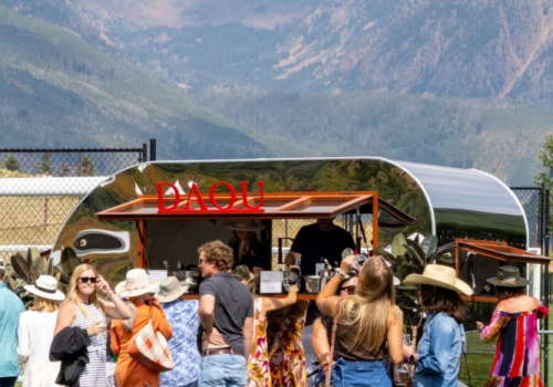 A sunny outdoor festival with a crowd near a food truck or bar, mountains in the background, people chatting and wearing casual summer outfits.
