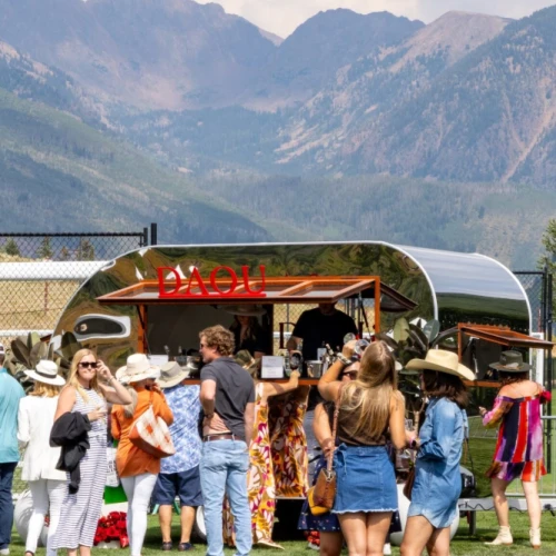 A sunny outdoor festival with a crowd near a food truck or bar, mountains in the background, people chatting and wearing casual summer outfits.
