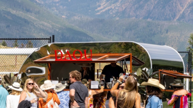 A sunny outdoor festival with a crowd near a food truck or bar, mountains in the background, people chatting and wearing casual summer outfits.