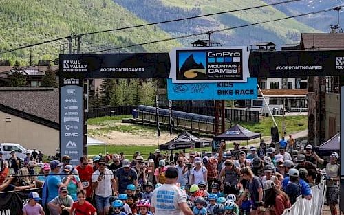 A crowded outdoor bike race finish area with participants, organizers, and mountains in the background, banners overhead, and spectators along the course.