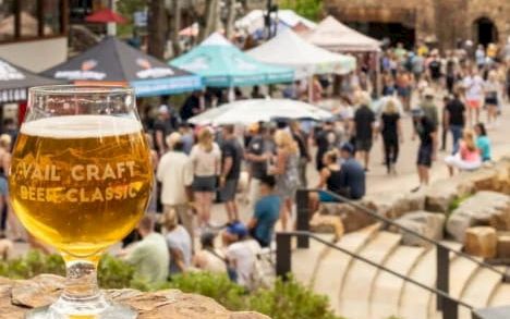 A glass of pale beer sits on a rock overlook at a crowded outdoor festival with tents and people in the background.