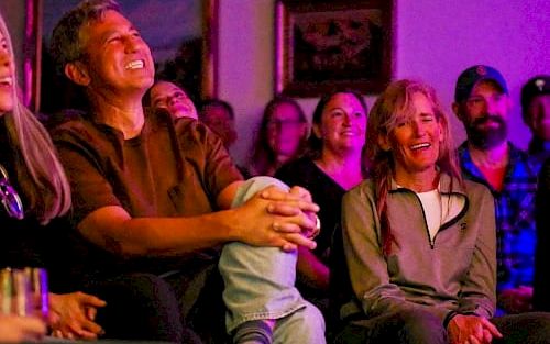 A group of people sitting close together, smiling and watching something entertaining in a cozy, dimly lit room.