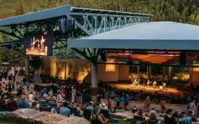 Shows an outdoor amphitheater or venue with a stage, audience seated on grass and benches, hillside backdrop, warm evening lighting. (ends with a period)