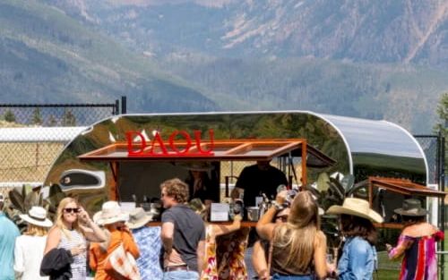 A group of people gathered around a mobile bar or food truck outdoors, with mountains in the background.