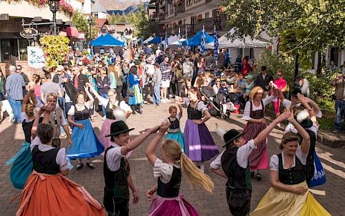 A lively outdoor festival on a sunny street with people dancing in colorful period costumes, market stalls, and a cheerful crowd.