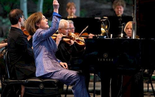 A conductor in a blue pinstripe suit raises a baton beside a grand piano and musicians in an outdoor orchestra, performing passionately.