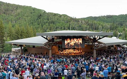 A large outdoor concert with a stage, crowd, and surrounding mountains; it&rsquo;s daytime and people are standing and seated on the grass.