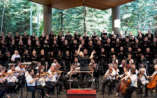 An outdoor orchestra and choir perform on a wooden stage with musicians playing violins, cellos, and brass, while a large choir fills the backdrop.