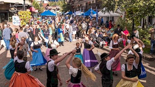 A lively street festival with colorful skirts, dancers, crowds, and market stalls lining a charming town street.