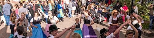 A lively street festival with colorful skirts, dancers, crowds, and market stalls lining a charming town street.