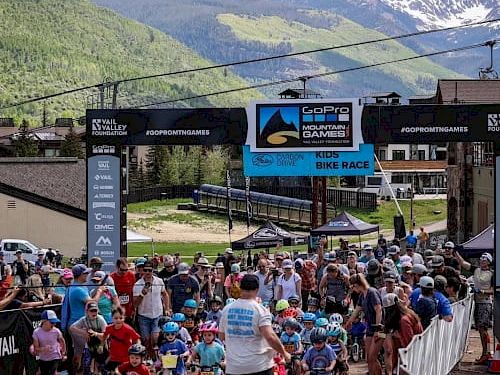 A crowd of cyclists and families gather at a mountain biking event start/finish, with banners and snowy peaks in the background.