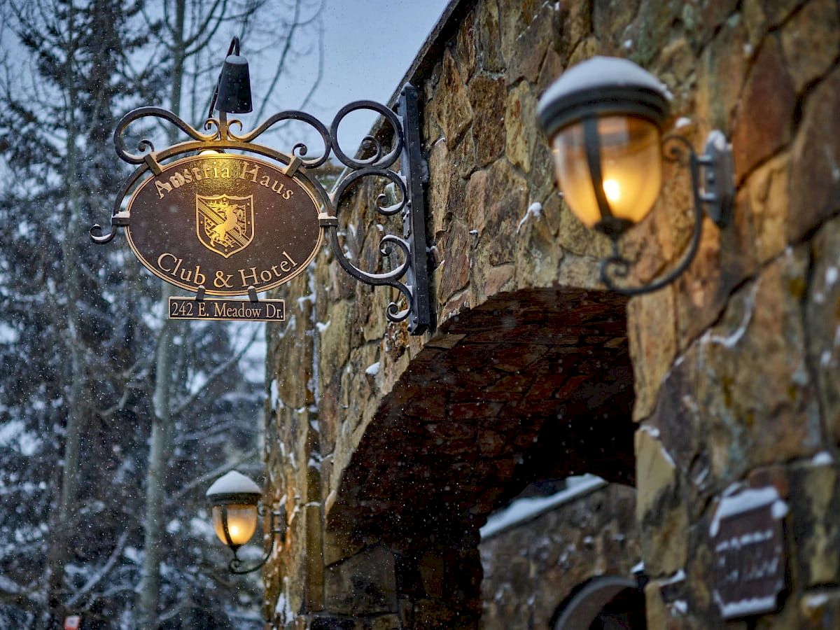 Snowy street outside a stone hotel with glowing lamps and a hanging crest sign for a club and hotel.