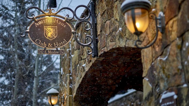 Snowy street outside a stone hotel with glowing lamps and a hanging crest sign for a club and hotel.