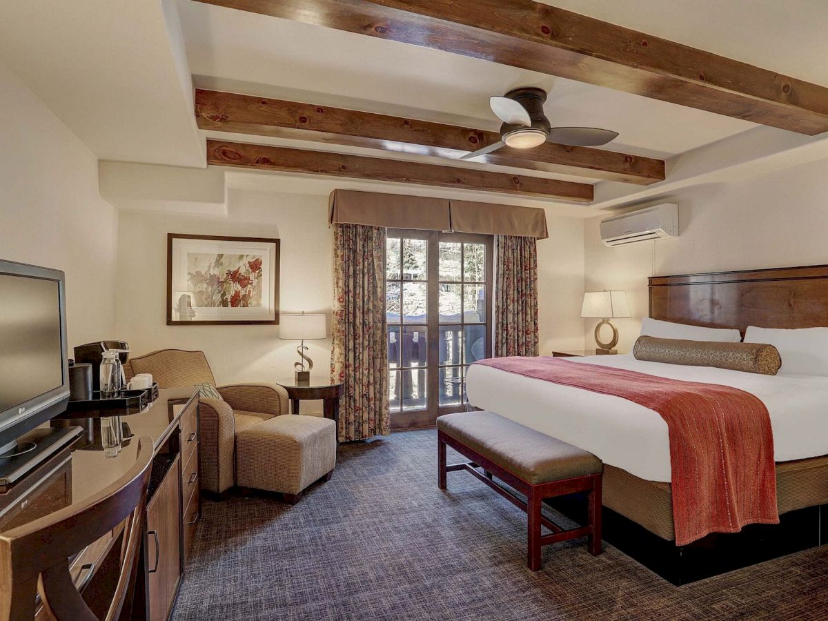 A hotel room with a king bed, wooden ceiling beams, a bench at the foot, sitting area with chairs, a desk, flat-screen TV, and a balcony door; cozy decor.