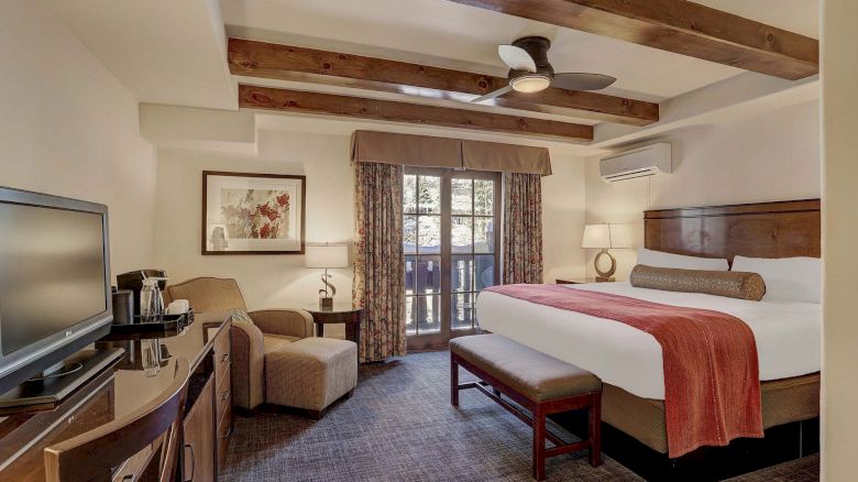 A hotel room with a king bed, wooden ceiling beams, a bench at the foot, sitting area with chairs, a desk, flat-screen TV, and a balcony door; cozy decor.