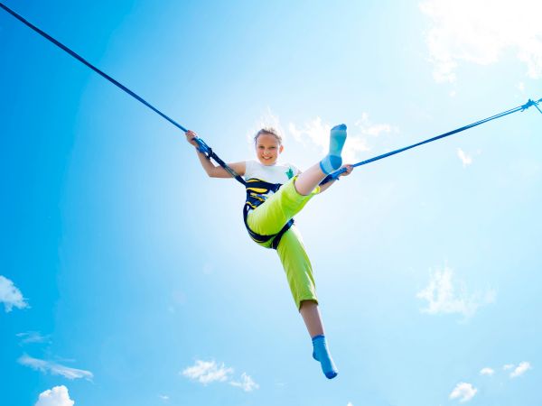 A smiling child is ziplining in the sky with arms out and a helmet harness, gliding above bright clouds.