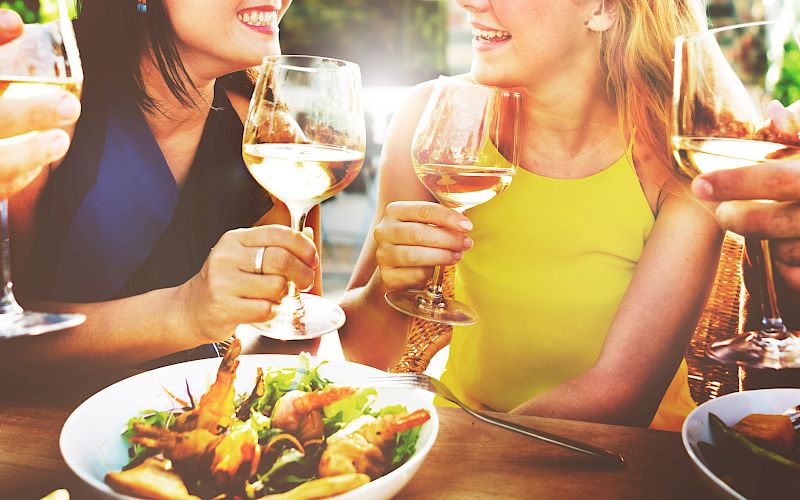 Friends dining outdoors, clinking glasses of white wine in sunshine, sharing smiles and good times together.
