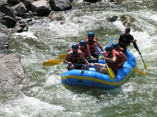 A group of people in helmets and life jackets white-water rafting on a blue inflatable raft through a rocky river gorge.