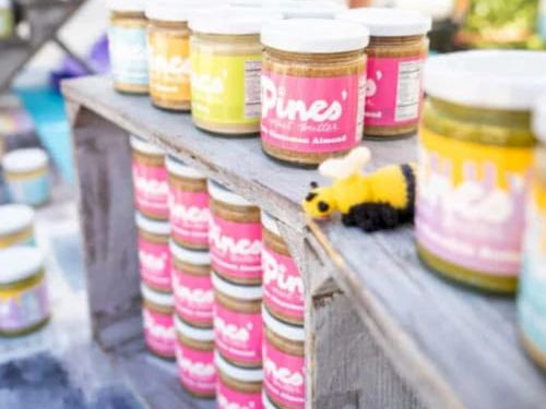 Jars of Pina&rsquo;s dip on a wooden table at a market, lined up with colorful labels and a tiny bee decoration in front.