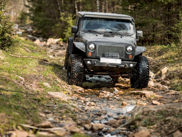 A rugged off-road Jeep splashes through a rocky creek in a forest, crawling over stones on huge tires.