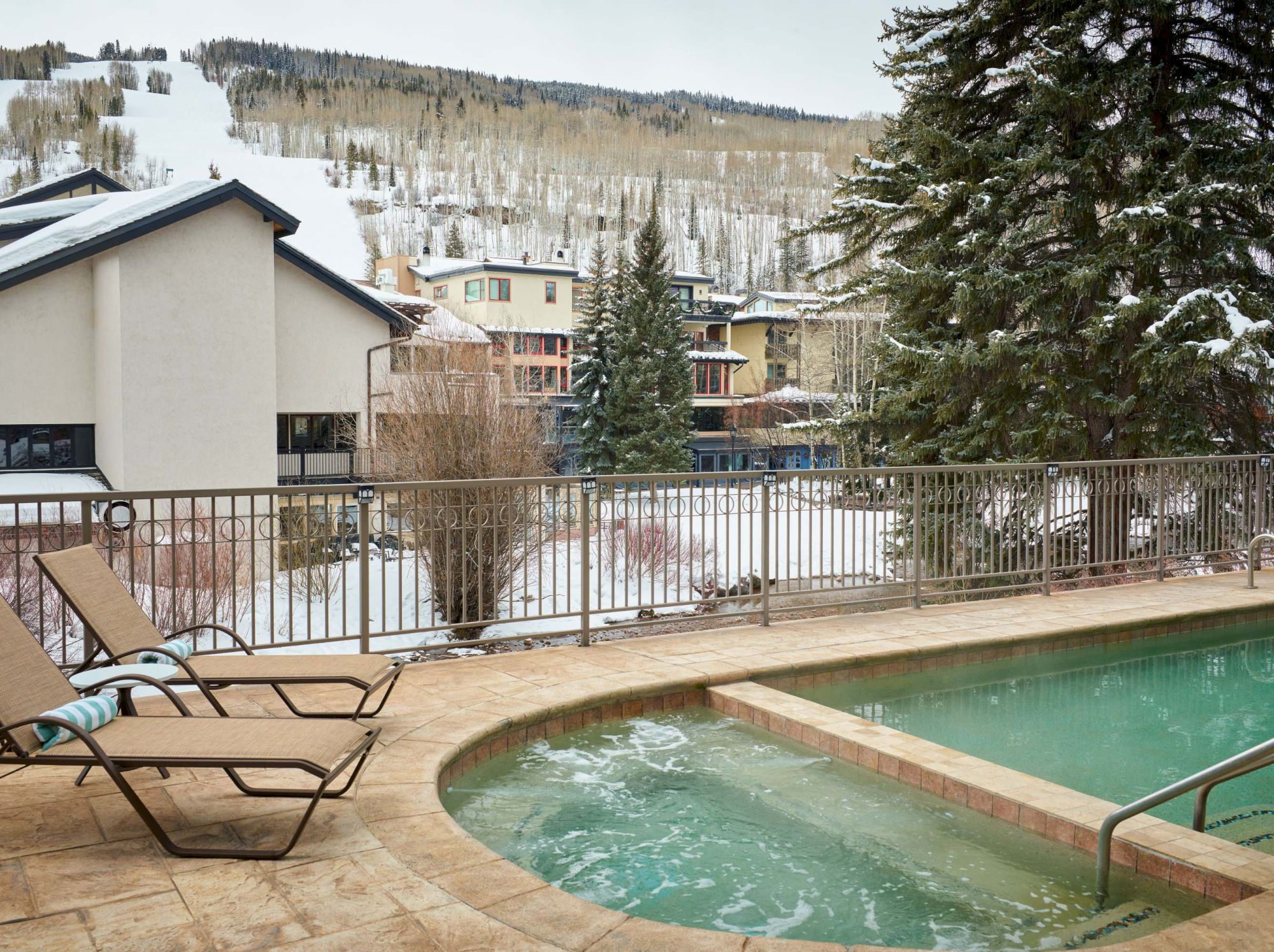 Snowy mountain resort view with an outdoor hot tub, lounge chairs, evergreen trees, and large apartment buildings.