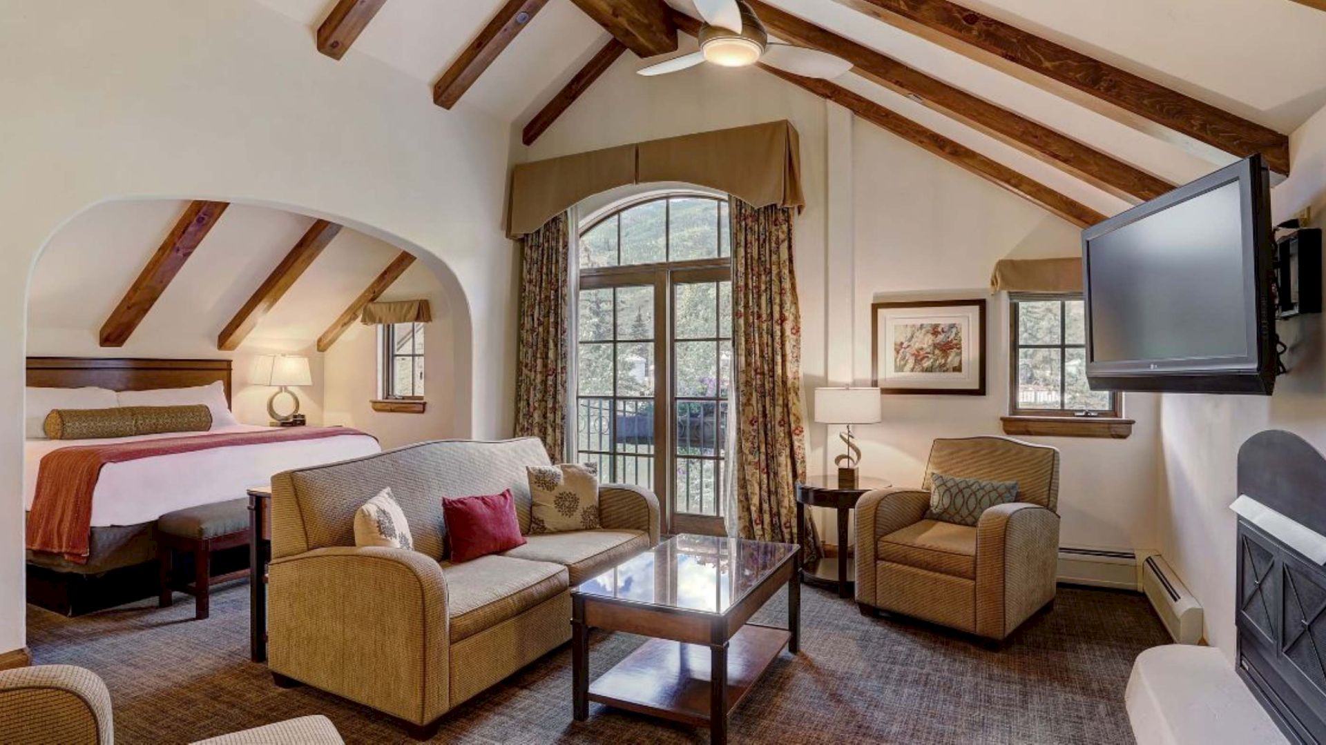 A cozy living room with vaulted wooden beams, fireplace and TV, cushioned chairs and couch, and a bed alcove by large windows.
