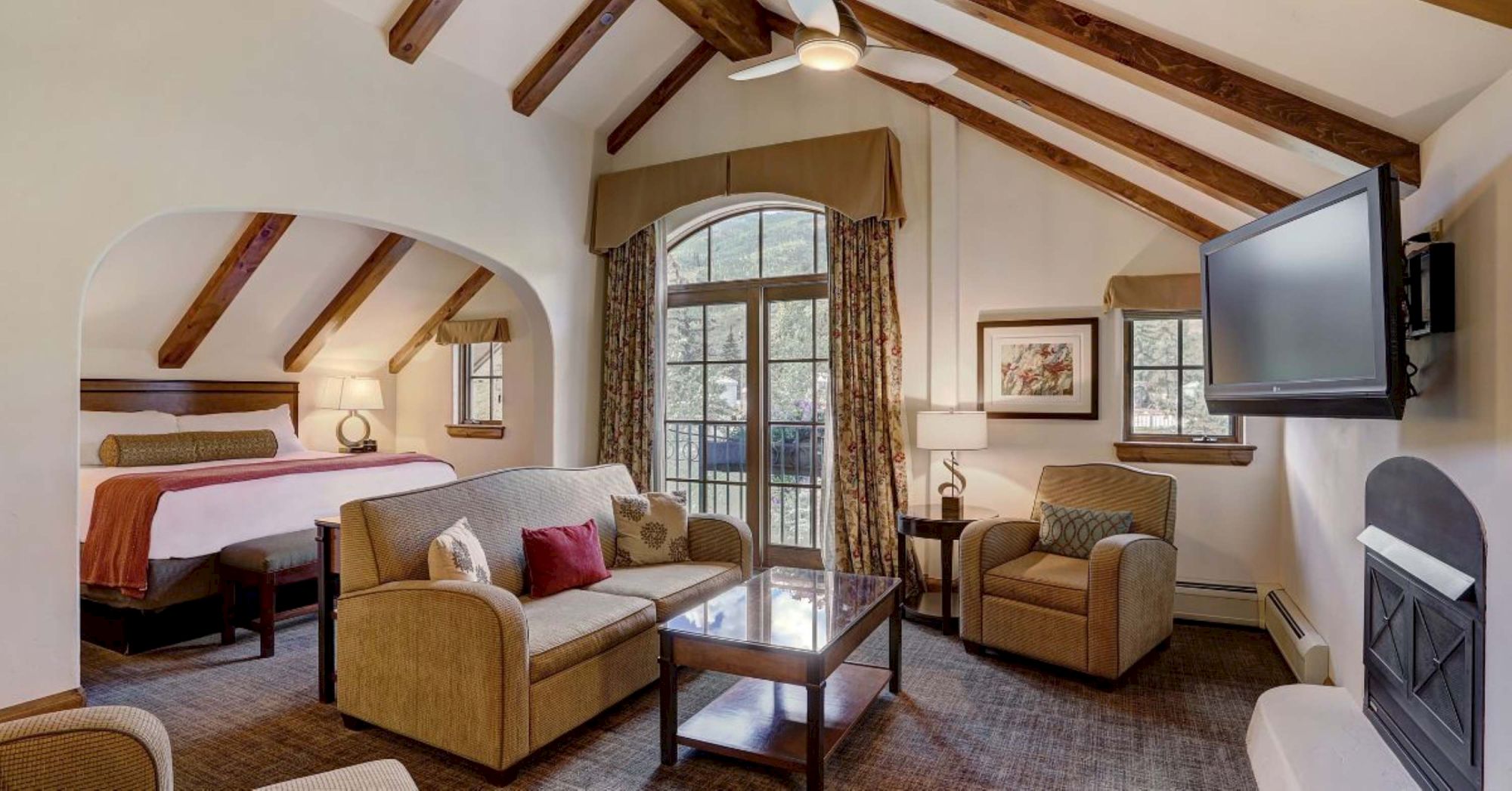 A cozy living room with vaulted wooden beams, fireplace and TV, cushioned chairs and couch, and a bed alcove by large windows.