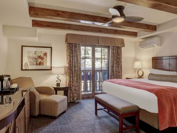Bright hotel bedroom with a king bed, patterned curtains, wood ceiling beams, ceiling fan, and a dresser with water glasses.